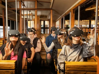 TimeRide SENSEUM - Virtual time travel through Cologne Sechs Personen in einem historischen Straßenbahnwagen, die Virtual-Reality-Headsets tragen und dabei in verschiedene Richtungen blicken. Die Passagiere wirken fasziniert und neugierig, während sie in eine virtuelle Welt eintauchen:Six people in a historic streetcar carriage wearing virtual reality headsets and looking in different directions. The passengers appear fascinated and curious as they immerse themselves in a virtual world: