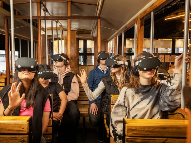 TimeRide SENSEUM - Virtuelle Zeitreise durch Köln Sechs Personen in einem historischen Straßenbahnwagen, die Virtual-Reality-Headsets tragen und dabei in verschiedene Richtungen blicken. Die Passagiere wirken fasziniert und neugierig, während sie in eine virtuelle Welt eintauchen: