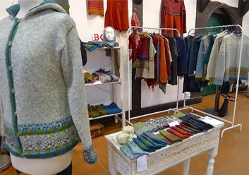 Eine bunte Auswahl handgefertigter Textilien, darunter Pullover und Schals, in einem Ausstellungsraum.