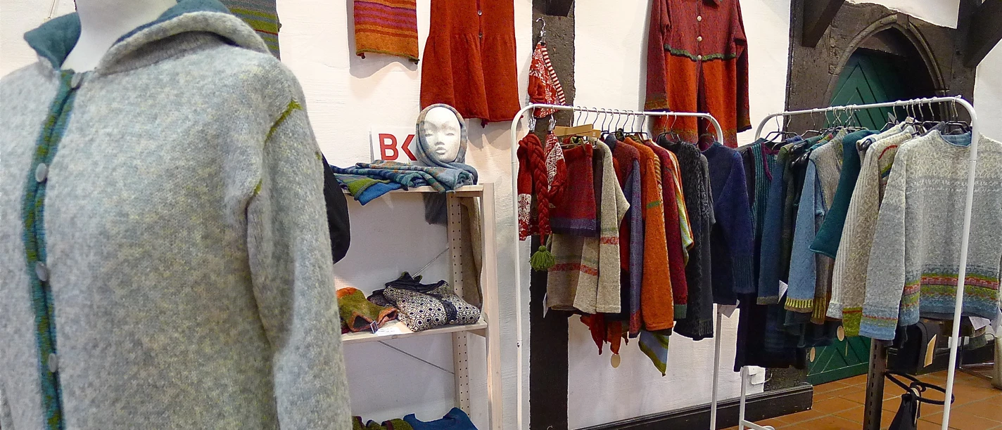 Eine bunte Auswahl handgefertigter Textilien, darunter Pullover und Schals, in einem Ausstellungsraum.
