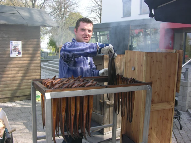 r-uchermarkt-aal-bruns Ein Mann in gestreiftem Hemd und mit Arbeitshandschuhen räuchert Aale in einem Holzofen im Freien.