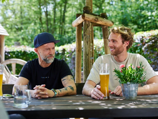 Holsteins Mühle Zwei Männer sitzen lächelnd an einem Holztisch im Freien mit Bier und einer Blumendekoration darauf.