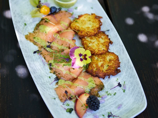 Holsteins Mühle Lachsfiletstreifen mit Kräutersoße, serviert mit Kartoffelpuffer und Obst, auf rustikalem Holztisch.