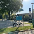 Fahrradständer unter Bäumen, leeres Parkfeld daneben, bei sonnigem Wetter, Ladestation sichtbar.