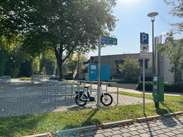 Fahrradständer unter Bäumen, leeres Parkfeld daneben, bei sonnigem Wetter, Ladestation sichtbar.