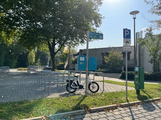 E-Bike Ladestation am Freibad Verl Fahrradständer unter Bäumen, leeres Parkfeld daneben, bei sonnigem Wetter, Ladestation sichtbar.
