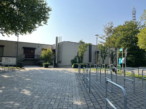 E-Bike Ladestation am Freibad Verl | Parkplatz Zum Meierhof Fahrradständer vor einem großen, flachen Gebäude mit Klinkerfassade und umgebender Bepflanzung.