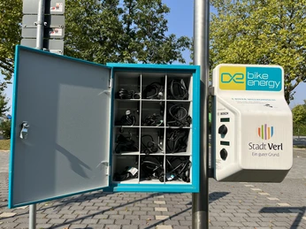 Kasten mit E-Bike Ladekabel an der Ladestation am Freibad Verl Barrierefreie E-Bike-Ladestation vor einem Stadtpark, ausgestattet mit mehreren Ladefächern und Anschlüssen.
