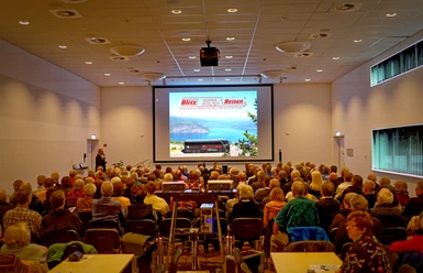 Blitz-Reisen Multivisionsshow-Norwegen Nordfjord Viele Menschen sitzen in einem Seminarraum und sehen eine Präsentation mit einem Bergsee-Bild an.