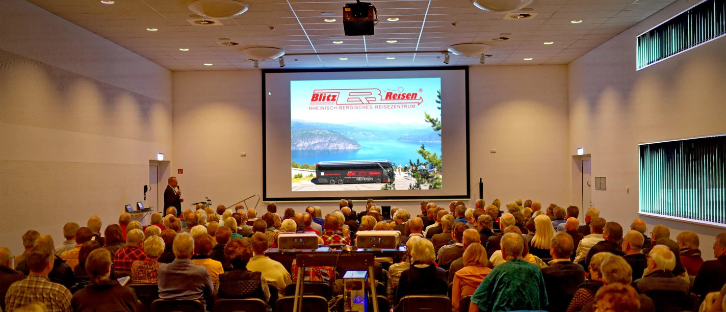 Blitz-Reisen Multivisionsshow-Norwegen Nordfjord Viele Menschen sitzen in einem Seminarraum und sehen eine Präsentation mit einem Bergsee-Bild an.