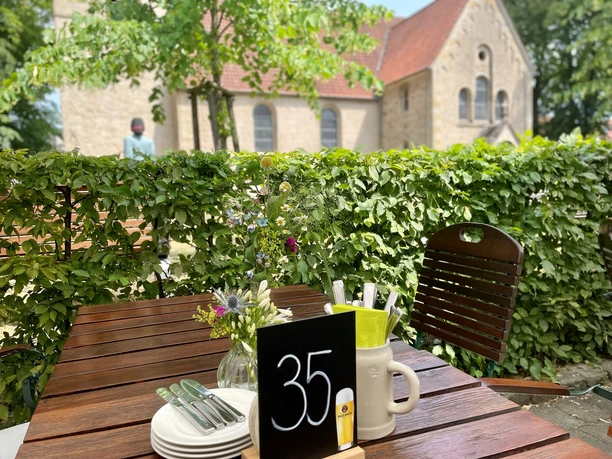 Emils Wirtshaus Terrassenansicht.jpg Holztisch unter freiem Himmel mit Blumen, Besteck und Tischnummer 35; Kirche im Hintergrund.