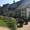 Emils Wirtshaus Terrassse.jpg Fachwerkhaus mit Terrasse, auf der Menschen gemütlich speisen, umgeben von gepflegten Pflanzen.