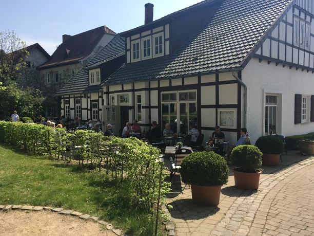 Emils Wirtshaus Terrassse.jpg Fachwerkhaus mit Terrasse, auf der Menschen gemütlich speisen, umgeben von gepflegten Pflanzen.