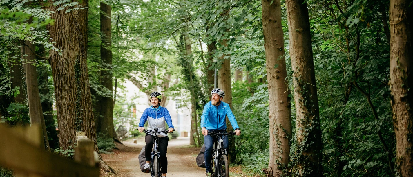 Brückenradweg Osnabrück - Bremen Two cyclists enjoy a ride on a forest path surrounded by tall trees.
