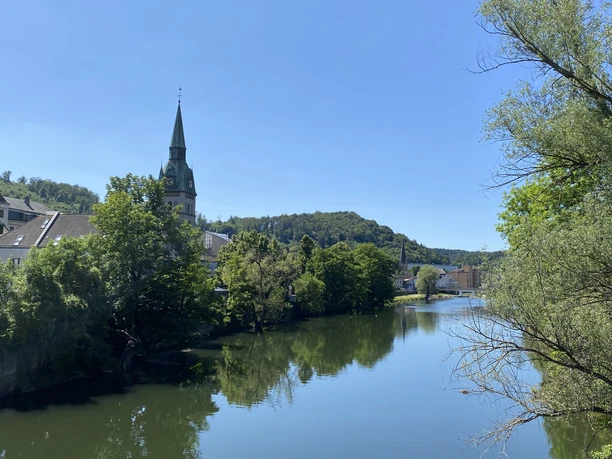 Eine friedliche Flusslandschaft mit einem Kirchturm im Zentrum, sanft eingebettet in grün bewaldete Hügel.
