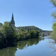 IMG_0905.jpg Eine friedliche Flusslandschaft mit einem Kirchturm im Zentrum, sanft eingebettet in grün bewaldete Hügel.