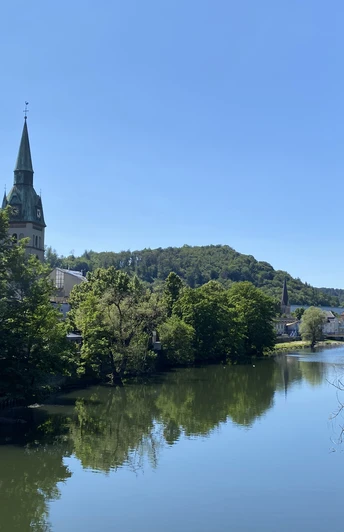 IMG_0905.jpg Eine friedliche Flusslandschaft mit einem Kirchturm im Zentrum, sanft eingebettet in grün bewaldete Hügel.