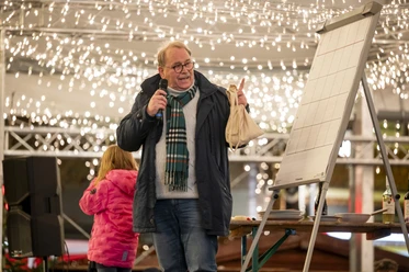Mann mit Mikrofon und Tasche bei Verlosung auf dem Weihnachtsmarkt unter festlicher Beleuchtung