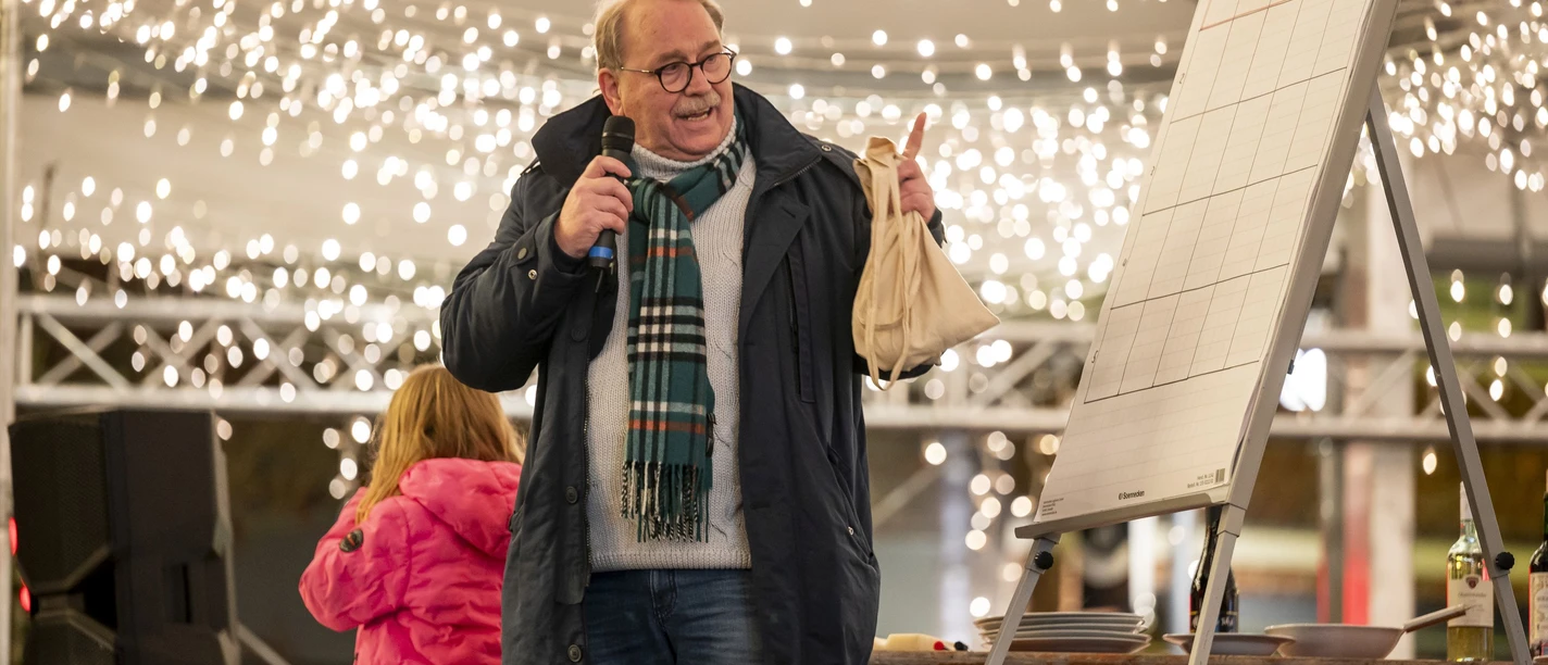 Goldene 7 Verlosung Weihnachtsmarkt Meppen s.jpg Mann mit Mikrofon und Tasche bei Verlosung auf dem Weihnachtsmarkt unter festlicher Beleuchtung