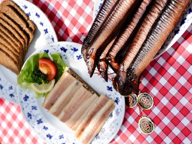 aale Auf einem rustikalen Tisch liegen auf einem karierten Tischtuch mehrere frisch geräucherte Aale kunstvoll nebeneinander auf einem Teller. Daneben befinden sich reich belegte Teller mit frischem Brot und zarten Heringsfilets, garniert mit Salat, Zitrone und einer Tomatenscheibe. Alles ist appetitlich auf weißen Tellern mit blauen Mustern angerichtet.