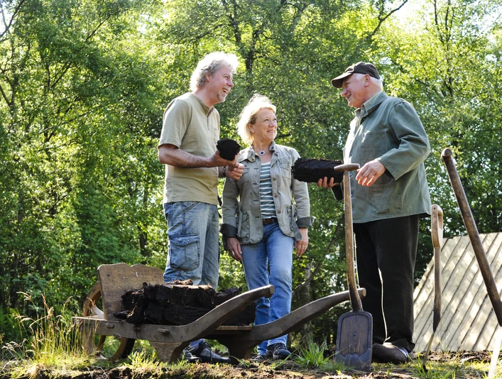 moorführung Drei Menschen stehen im Moor und unterhalten sich, während sie Torfstücke in den Händen halten. Eine Schubkarre voller Torf steht neben ihnen.