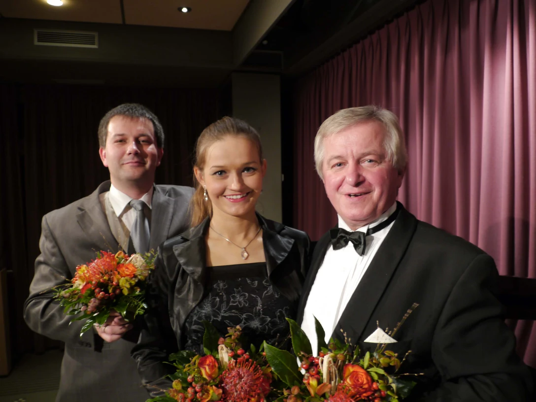 gala-oper-operette-musical Drei elegant gekleidete Personen mit Blumensträußen vor bordeauxrotem Bühnenvorhang.