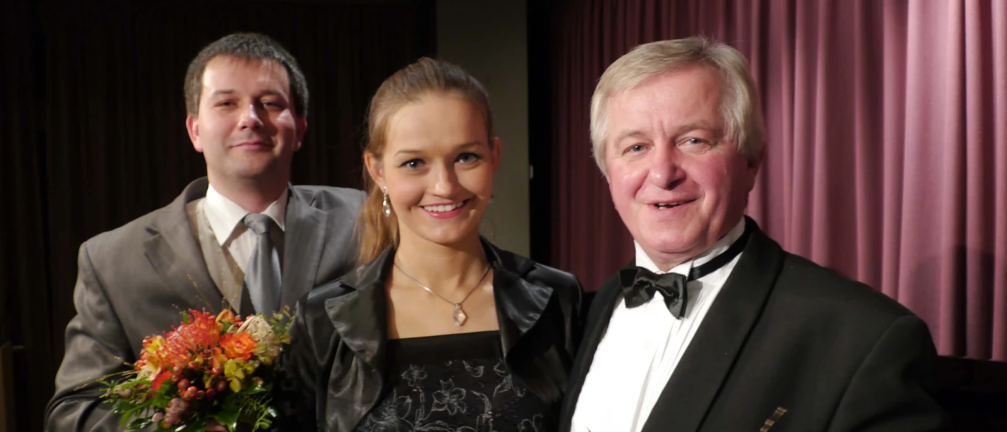 gala-oper-operette-musical Drei elegant gekleidete Personen mit Blumensträußen vor bordeauxrotem Bühnenvorhang.