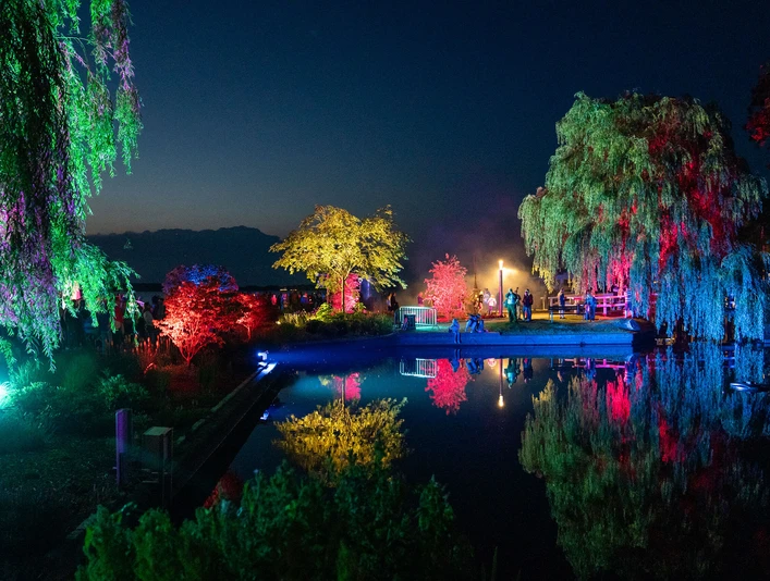 Lichternacht im Zwischenahner Kurpark Das Bild zeigt eine nächtliche Szene im Zwischenahner Kurpark, wo buntes Licht Bäume und Wasser in lebendige Farben taucht. Die Reflexionen erzeugen eine faszinierende Symphonie aus leuchtendem Rot, Blau und Grün auf dem ruhigen Teich, während die Silhouetten von Besuchern die Szenerie beleben.The picture shows a nocturnal scene in the Zwischenahn spa gardens, where colorful light bathes trees and water in vibrant colors. The reflections create a fascinating symphony of bright red, blue and green on the calm pond, while the silhouettes of visitors enliven the scenery.Billedet viser en natlig scene i Zwischenahns spahave, hvor det skarpe lys bader træerne og vandet i levende farver. Refleksionerne skaber en fascinerende symfoni af lyse rødt, blåt og grønt i den rolige dam, mens silhuetterne af de besøgende giver liv til sceneriet.De foto toont een nachtelijk tafereel in het kuurpark Zwischenahn, waar helder licht de bomen en het water in levendige kleuren hult. De weerspiegelingen creëren een fascinerende symfonie van helder rood, blauw en groen op de kalme vijver, terwijl de silhouetten van bezoekers het landschap verlevendigen.