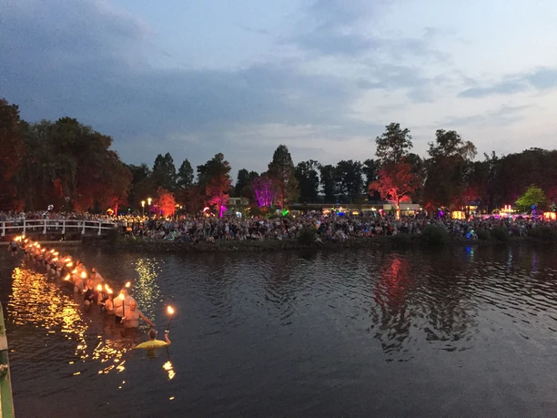 fackelschwimmen Fackelschwimmen bei der Dämmerung: Menschenmengen säumen das Ufer, während glühende Fackeln auf dem Wasser treiben und eine festliche Atmosphäre entsteht.