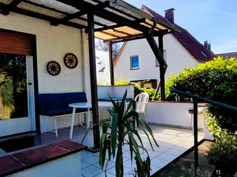 Haus am Park 3 (C) Gaby Schramm.jpg Überdachte Terrasse mit Tisch, Stühlen und Pflanzen, umgeben von Büschen und angrenzenden Gebäuden.