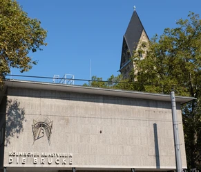 Kölnischer Kunstverein Der Kölnische Kunstverein: Moderne Betonarchitektur vor einem Kirchturmhintergrund, von Bäumen umrahmt.