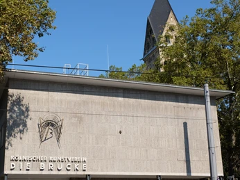Kölnischer Kunstverein Der Kölnische Kunstverein: Moderne Betonarchitektur vor einem Kirchturmhintergrund, von Bäumen umrahmt.The Kölnischer Kunstverein: Modern concrete architecture in front of a church tower background, framed by trees.