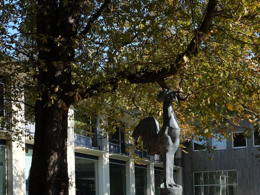 Kölnischer Kunstverein Ein bronzeener Hahn thront auf einem gemauerten Sockel vor einem modernen Gebäude, umgeben von Herbstlaub.A bronze cockerel is enthroned on a brick plinth in front of a modern building, surrounded by autumn leaves.