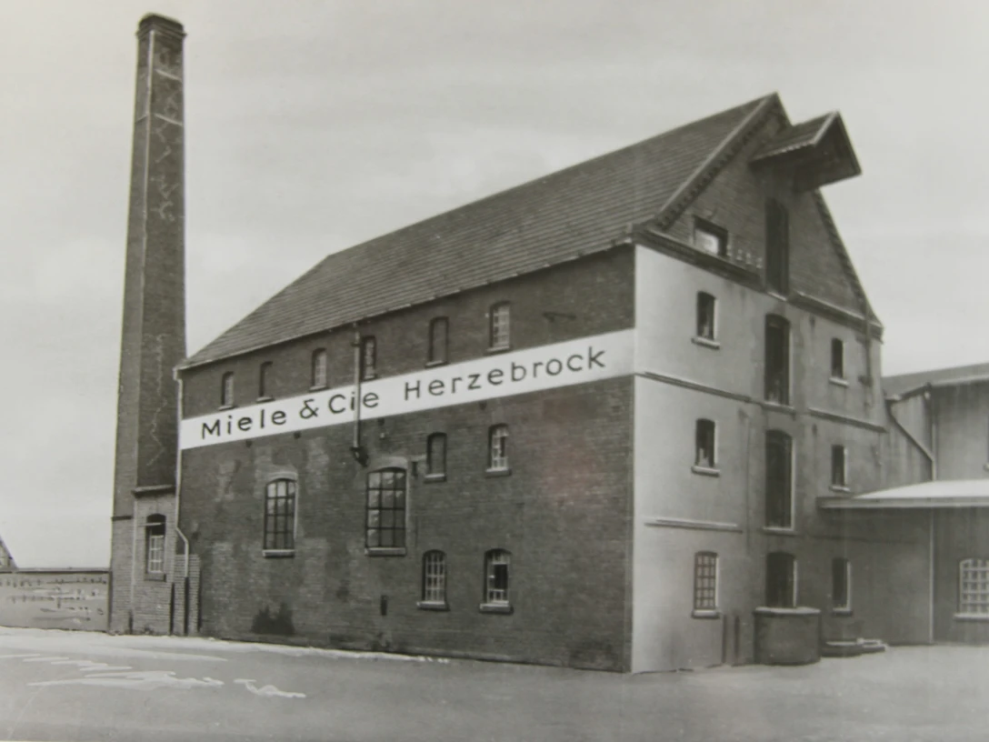 Ehemalige Nöllmann’sche Dampf, Mahl- und Sägemühle Ein historisches Backsteingebäude mit "Miele & Cie Herzebrock"-Schriftzug und hohem Schornstein.