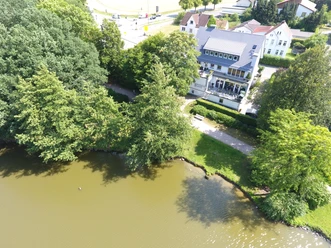 Restaurant am Seeufer, umgeben von Bäumen. Sitzplätze auf Terrasse, Zugang über gepflasterten Weg.