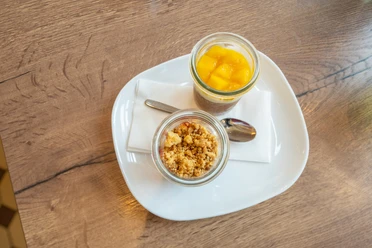 Café Bös Frühstücksteller, Ratingen Zwei Gläser auf einem weißen Teller, gefüllt mit frischem Obst und knusprigem Crumble, präsentiert.