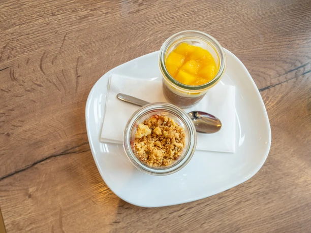 Café Bös Frühstücksteller, Ratingen Zwei Gläser auf einem weißen Teller, gefüllt mit frischem Obst und knusprigem Crumble, präsentiert.