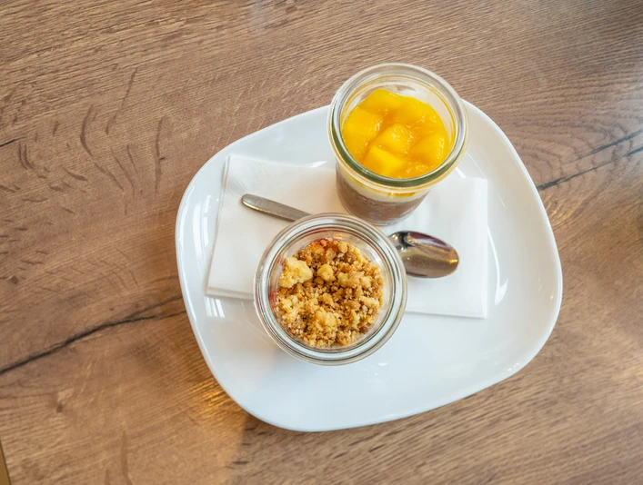Café Bös Frühstücksteller, Ratingen Zwei Gläser auf einem weißen Teller, gefüllt mit frischem Obst und knusprigem Crumble, präsentiert.