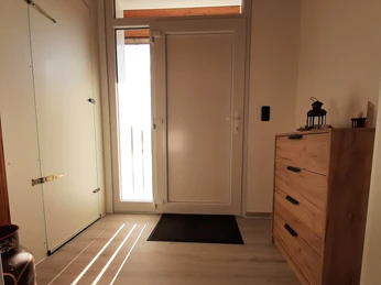 20240825_173503.jpg Heller Hausflur mit Holzboden, links eine Tür mit Glaselementen, rechts eine Holzkommode. Lichtstrahlen fallen herein.