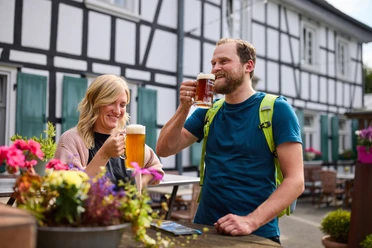 Ein lächelndes Paar genießt Bier vor einem Fachwerkhaus mit blühenden Pflanzen im Vordergrund.