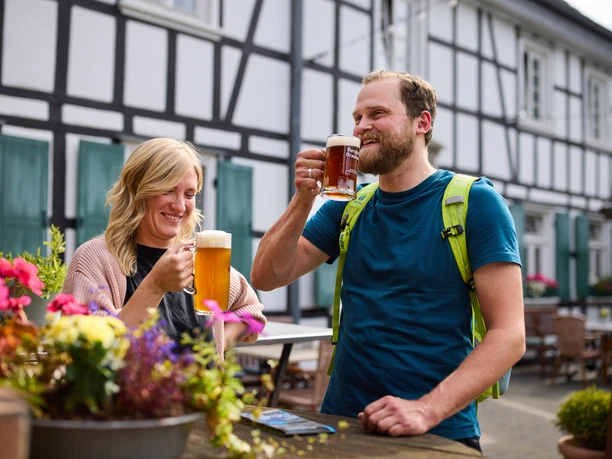 Restaurant Zur Post Ein lächelndes Paar genießt Bier vor einem Fachwerkhaus mit blühenden Pflanzen im Vordergrund.