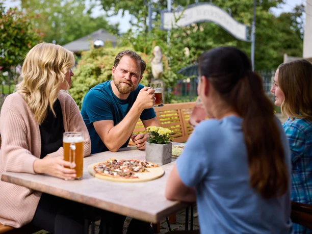 Restaurant Zur Post <p>Vier Personen genießen Getränke und Pizza in einem sonnigen Biergarten, umgeben von grüner Natur.</p>