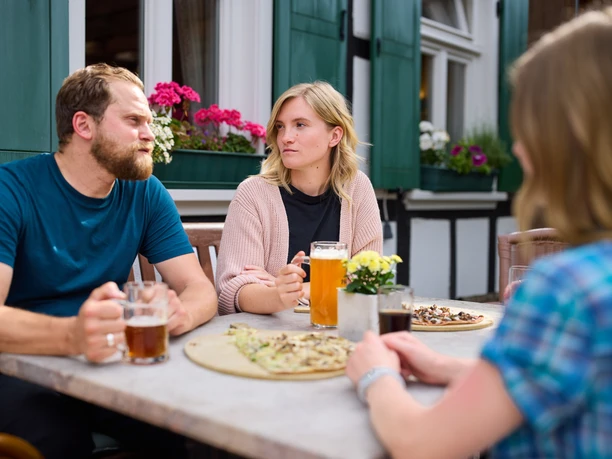 Restaurant Zur Post Drei Personen genießen Bier und Flammkuchen an einem Tisch im Freien eines gemütlichen Restaurants.