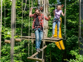 Waldkletterpark Velbert Menschen mit Sicherheitsgurten navigieren über Hängebrücken zwischen Bäumen in einem Kletterpark.