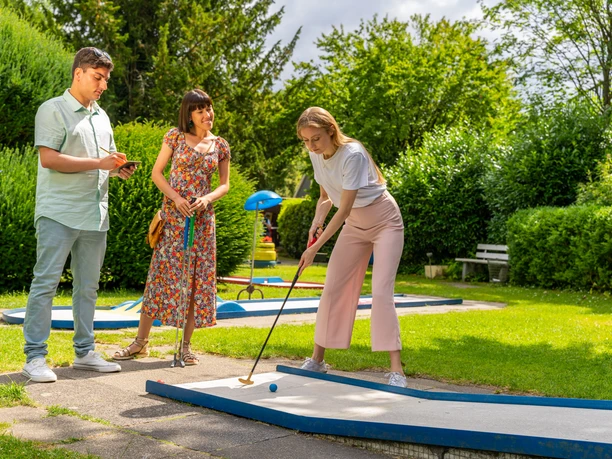 Minigolf spielen in Velbert Drei Personen spielen Minigolf auf einer Anlage im Grünen, umgeben von Bäumen und Hecken.