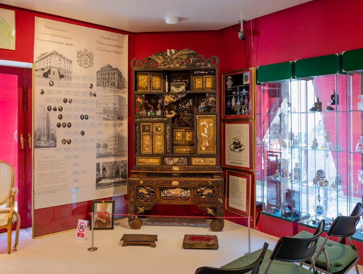 Duftmuseum Farina Ein opulent dekorierter Raum im Farina Duftmuseum zeigt einen reich verzierten Barockschrank mit bunten Parfümflaschen in einer gläsernen Vitrine, hell beleuchtet und eingerahmt von roten Wänden.An opulently decorated room in the Farina Fragrance Museum shows a richly decorated baroque cabinet with colorful perfume bottles in a glass display case, brightly lit and framed by red walls.