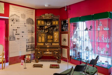 Farina Fragrance Museum An opulently decorated room in the Farina Fragrance Museum shows a richly decorated baroque cabinet with colorful perfume bottles in a glass display case, brightly lit and framed by red walls.