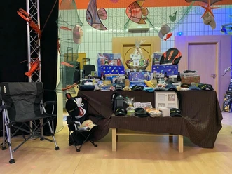 „Bunt dekorierter Bühnenbereich mit Netz‑ und Fischausschmückung, davor ein Tisch mit braunem Tischtuch, auf dem zahlreiche kleine Geschenkpakete, Korbgeschenke, Sport‑ und Outdoor‑Artikel wie Rucksack und Caps arrangiert sind. Neben dem Tisch stehen zwei Campingstühle, in dem einen ein Stofftier und weiterer kleiner Krimskrams.“