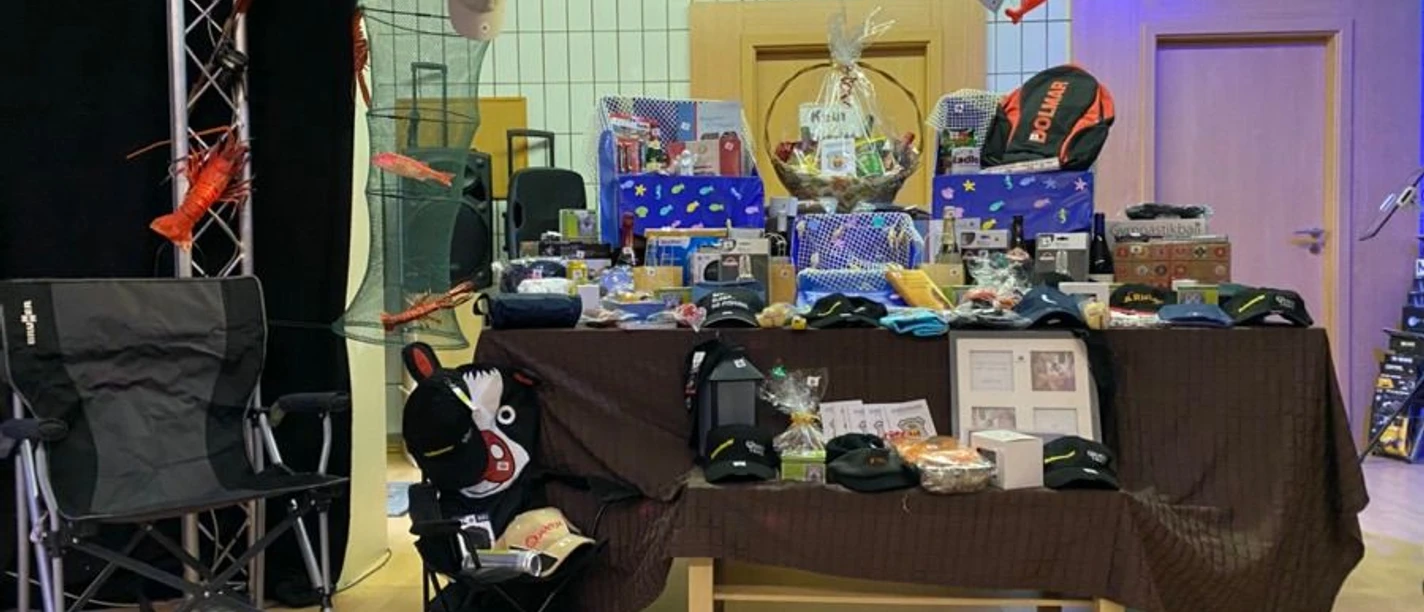 „Bunt dekorierter Bühnenbereich mit Netz‑ und Fischausschmückung, davor ein Tisch mit braunem Tischtuch, auf dem zahlreiche kleine Geschenkpakete, Korbgeschenke, Sport‑ und Outdoor‑Artikel wie Rucksack und Caps arrangiert sind. Neben dem Tisch stehen zwei Campingstühle, in dem einen ein Stofftier und weiterer kleiner Krimskrams.“