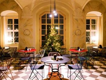 Gemütlicher Raum mit weihnachtlich geschmücktem Baum, runden Tischen und bunten Stühlen auf einem Schachbrettboden, warm beleuchtet.Cozy room with a tree decorated for Christmas, round tables and colorful chairs on a chessboard floor, warmly lit.Útulná místnost s vánočně ozdobeným stromečkem, kulatými stoly a barevnými židlemi na šachovnicové podlaze, příjemně osvětlená.Przytulna sala z choinką udekorowaną na Boże Narodzenie, okrągłymi stolikami i kolorowymi krzesłami na podłodze w szachownicę, ciepło oświetlona.Gezellige ruimte met een kerstboom, ronde tafels en kleurrijke stoelen op een schaakbordvloer, warm verlicht.Sala accogliente con albero addobbato per Natale, tavoli rotondi e sedie colorate su un pavimento a scacchiera, illuminato con luce calda.